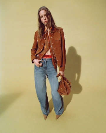 Model in brown suede jacket and blue jeans holding a handbag against a yellow studio backdrop