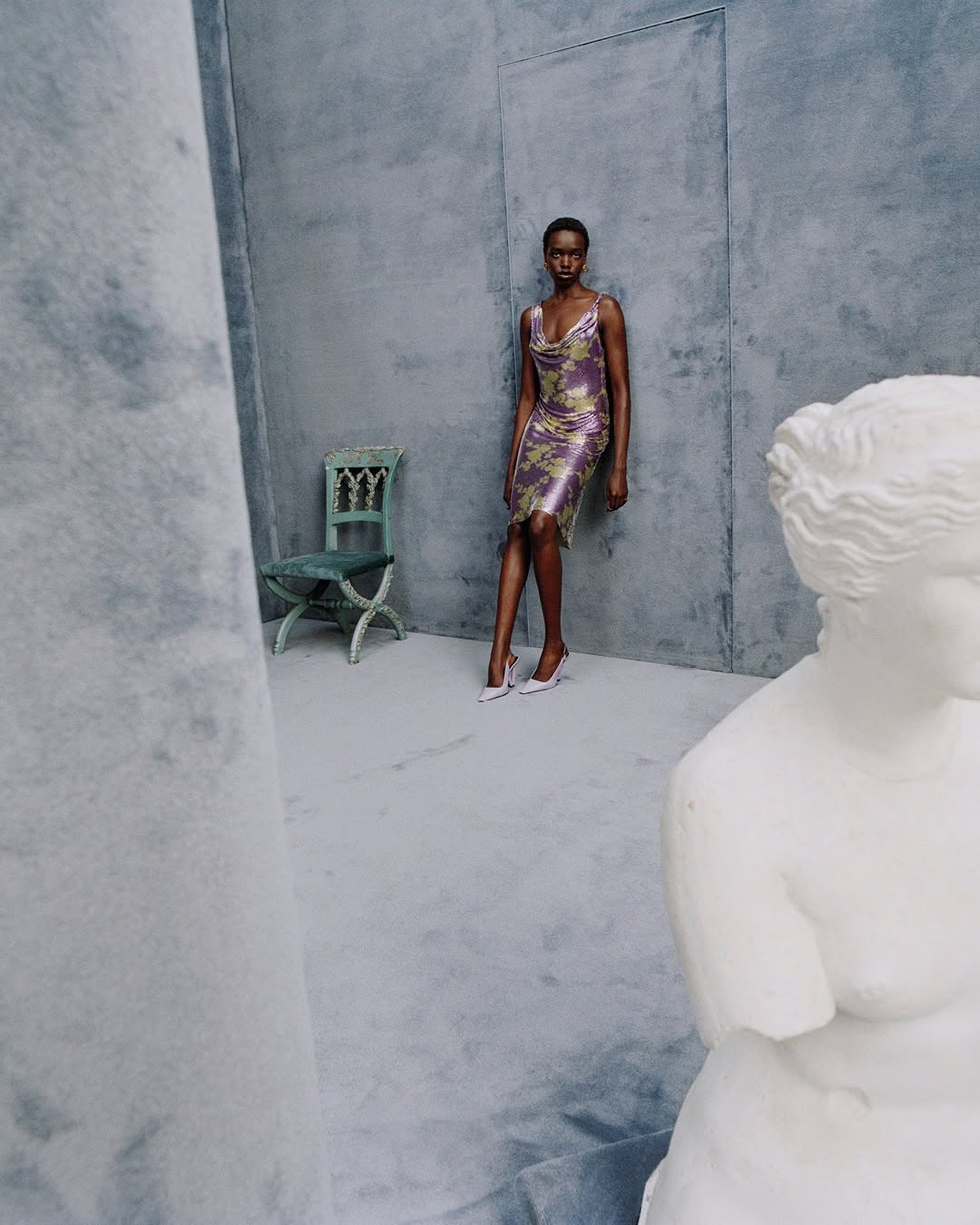 Model in a patterned purple dress leaning against a grey wall next to a chair and a white marble bust.