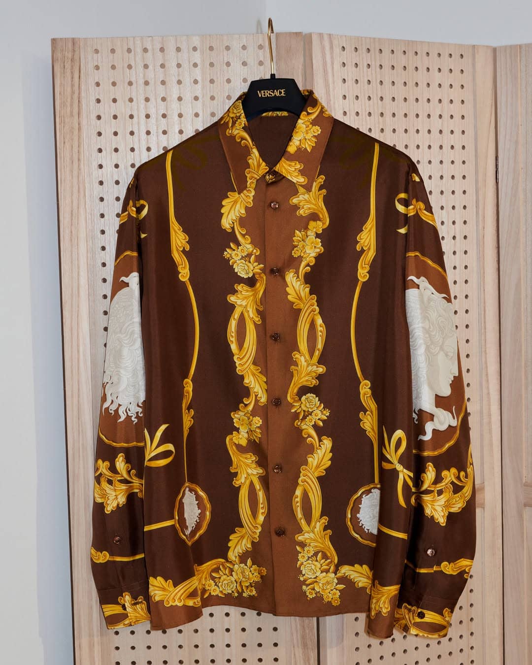 Brown long-sleeve silk shirt with intricate gold baroque print hanging on a black branded hanger against a pegboard wall