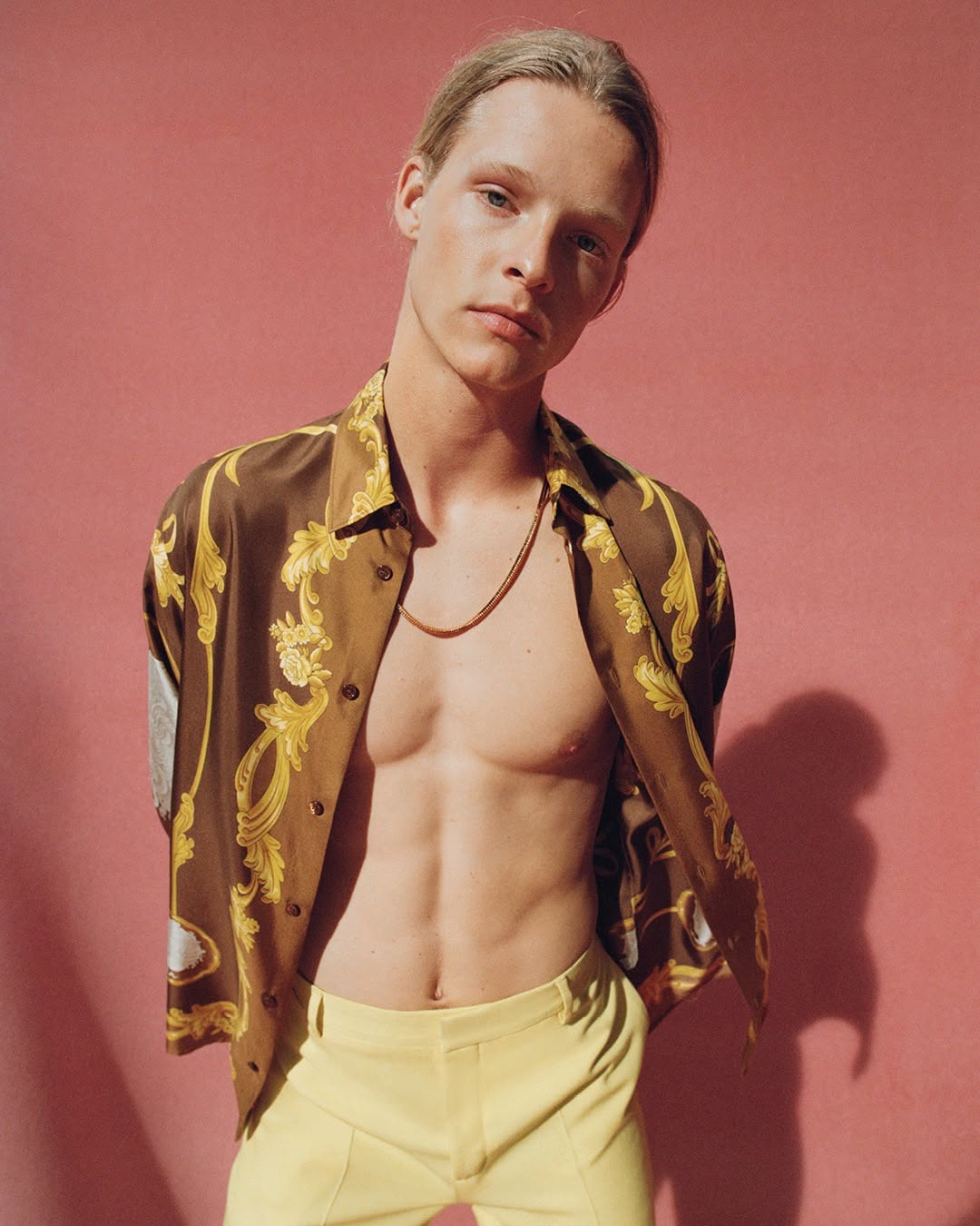 Blonde male model wearing an unbuttoned brown patterned silk shirt and yellow trousers against a solid pink backdrop