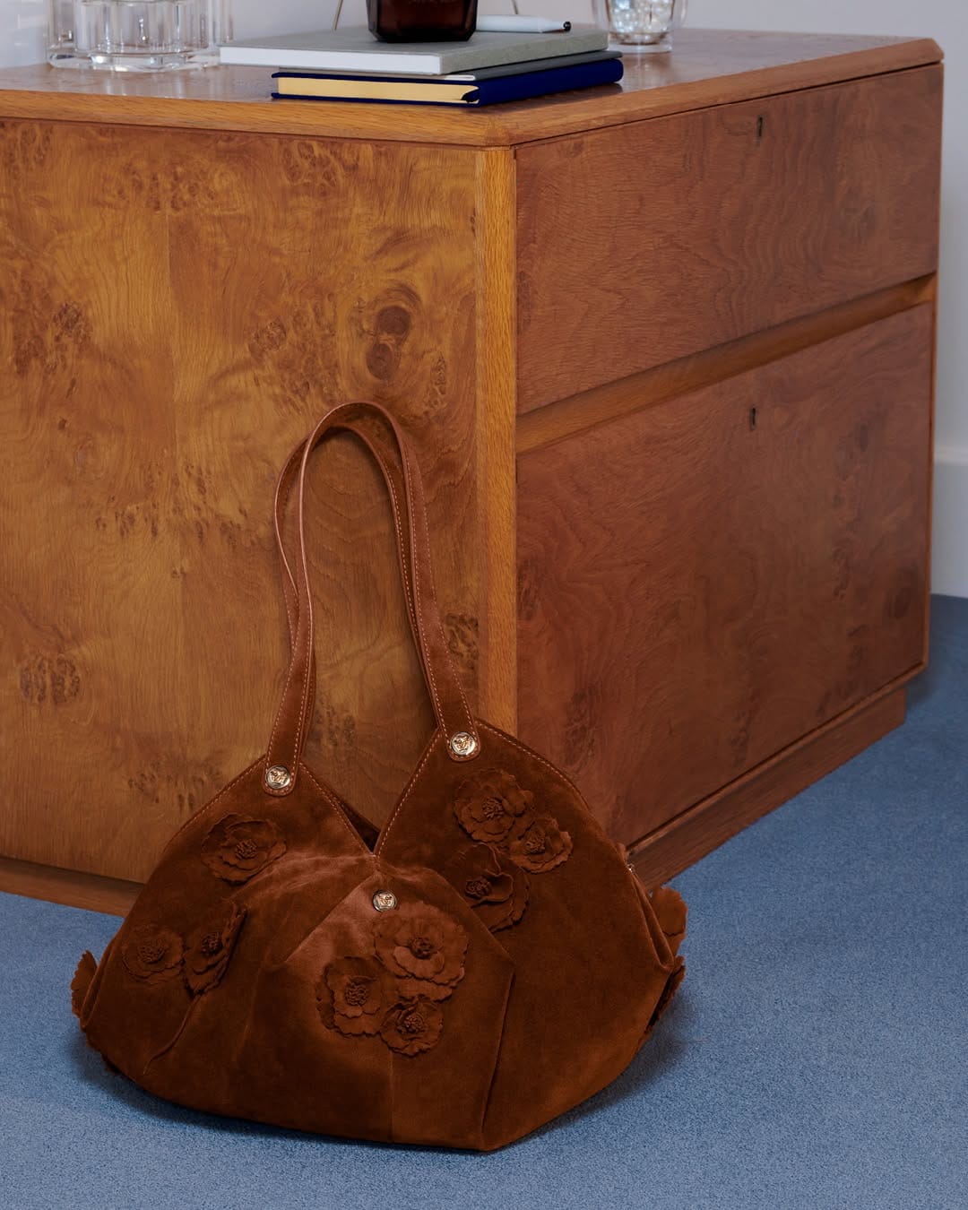 Brown leather handbag with floral appliques resting on a blue floor in front of a wooden cabinet.