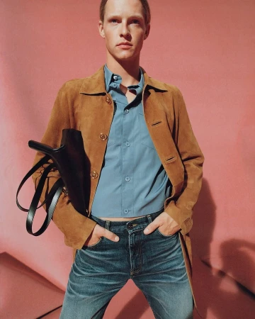 Male model in a brown suede jacket, blue shirt and jeans holding a black handbag against a pink background