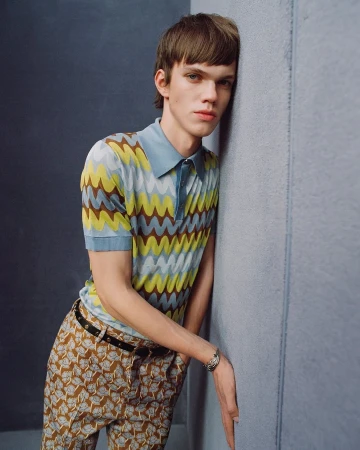 Young male model leaning against a grey wall wearing a patterned polo shirt and patterned trousers for Versace