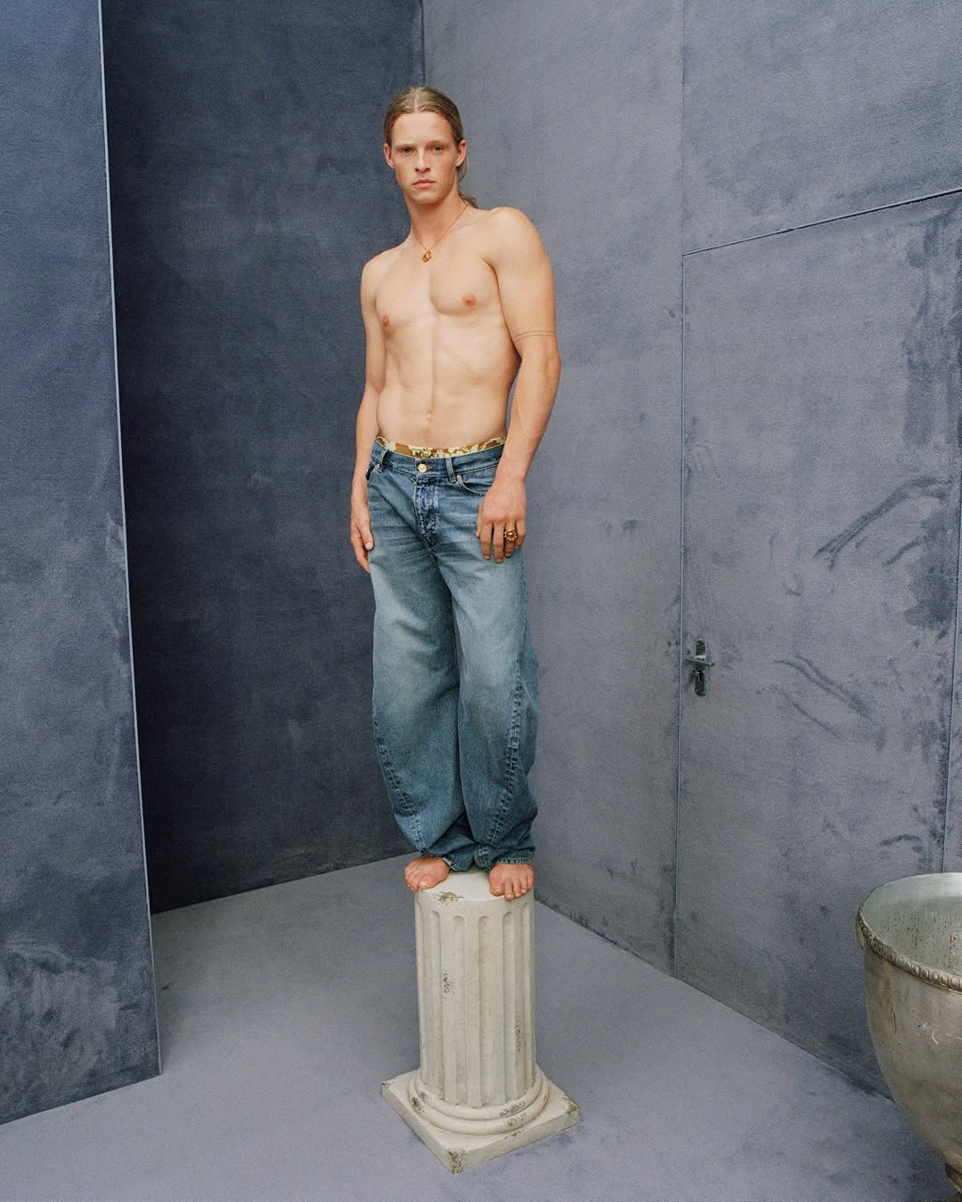 Shirtless male model standing barefoot on a small stone pillar wearing blue denim jeans against a textured grey wall background