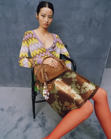 Model seated on a black chair holding a brown leather handbag, wearing a patterned cardigan and metallic skirt against a grey textured studio backdrop.