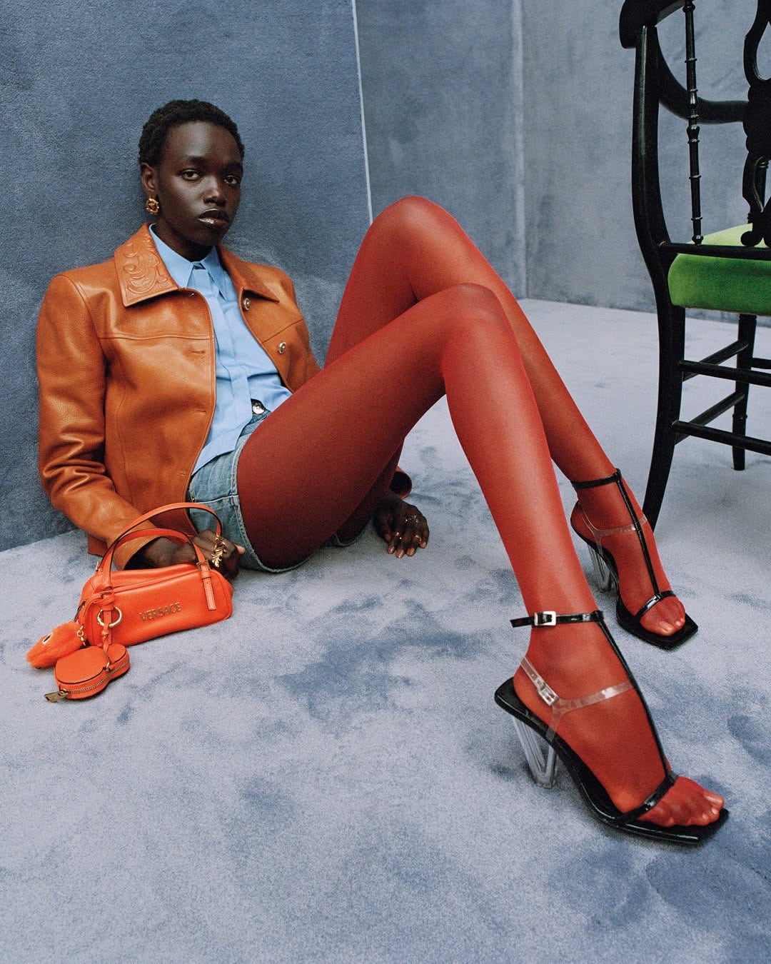 Model seated on grey studio floor wearing a brown leather jacket and orange tights, next to an orange handbag.