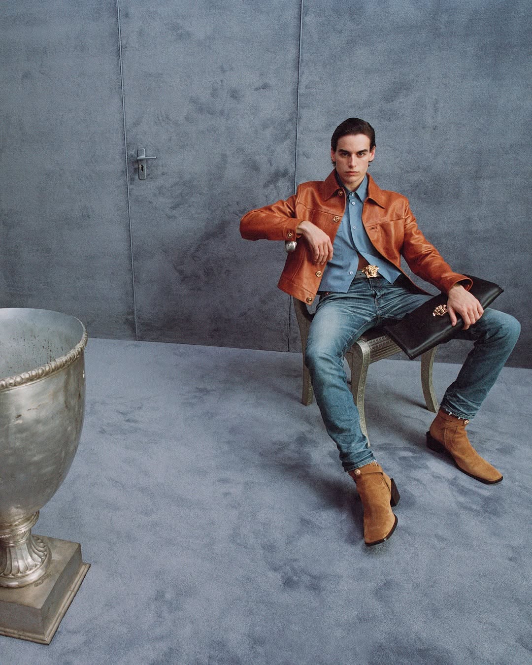 Man seated on a chair wearing a brown leather jacket and blue jeans against a textured grey studio background