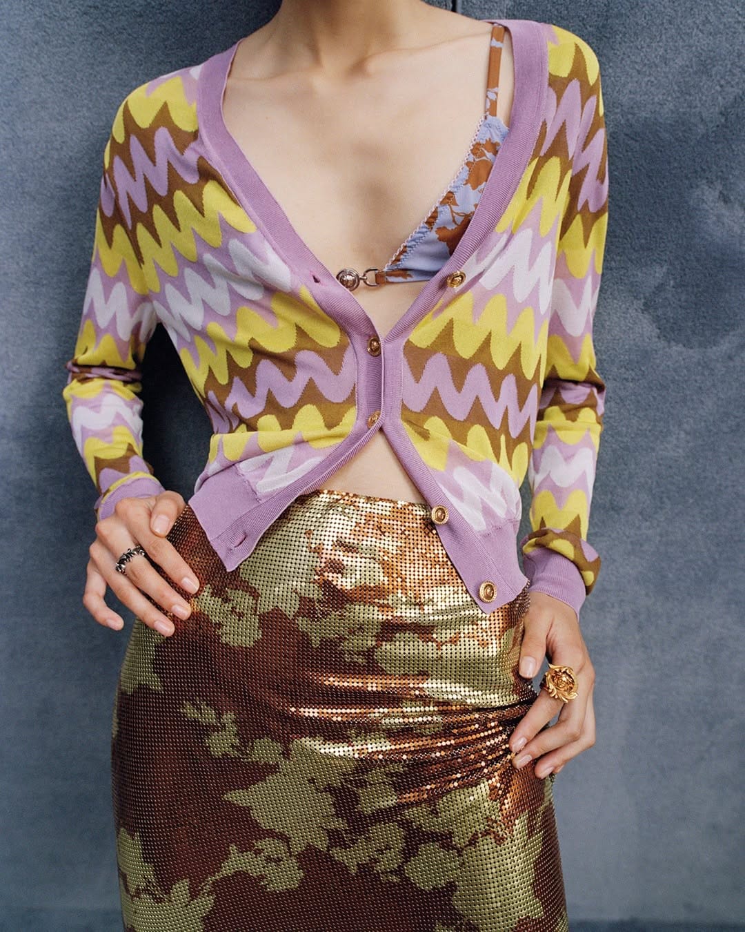 Model wearing a chevron-patterned cardigan and metallic sequin skirt standing before a textured grey wall for Versace.
