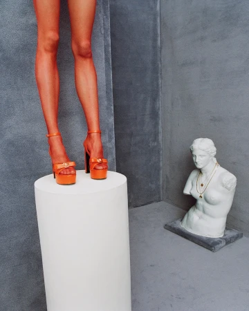 Model standing on a white pedestal wearing platform heels with a classical bust in the background for Versace Spring 2025 campaign