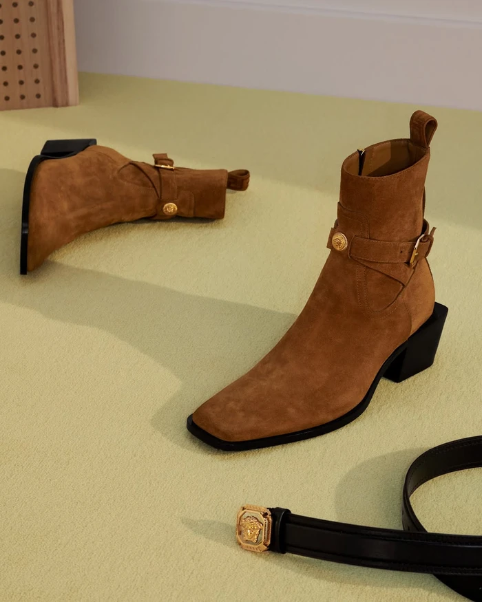 Tan suede ankle boots with buckle straps and black block heels resting on a pale green surface.