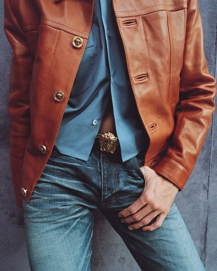 Model wearing a brown leather jacket, blue button-down shirt, and denim jeans with a gold belt buckle