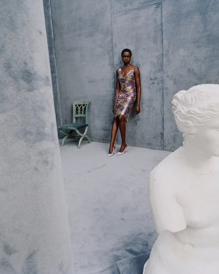 Model in a patterned purple dress leaning against a grey wall next to a chair and a white marble bust.