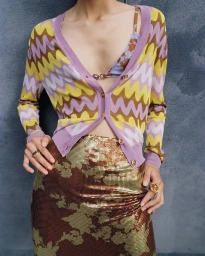 Model wearing a chevron-patterned cardigan and metallic sequin skirt standing before a textured grey wall for Versace.