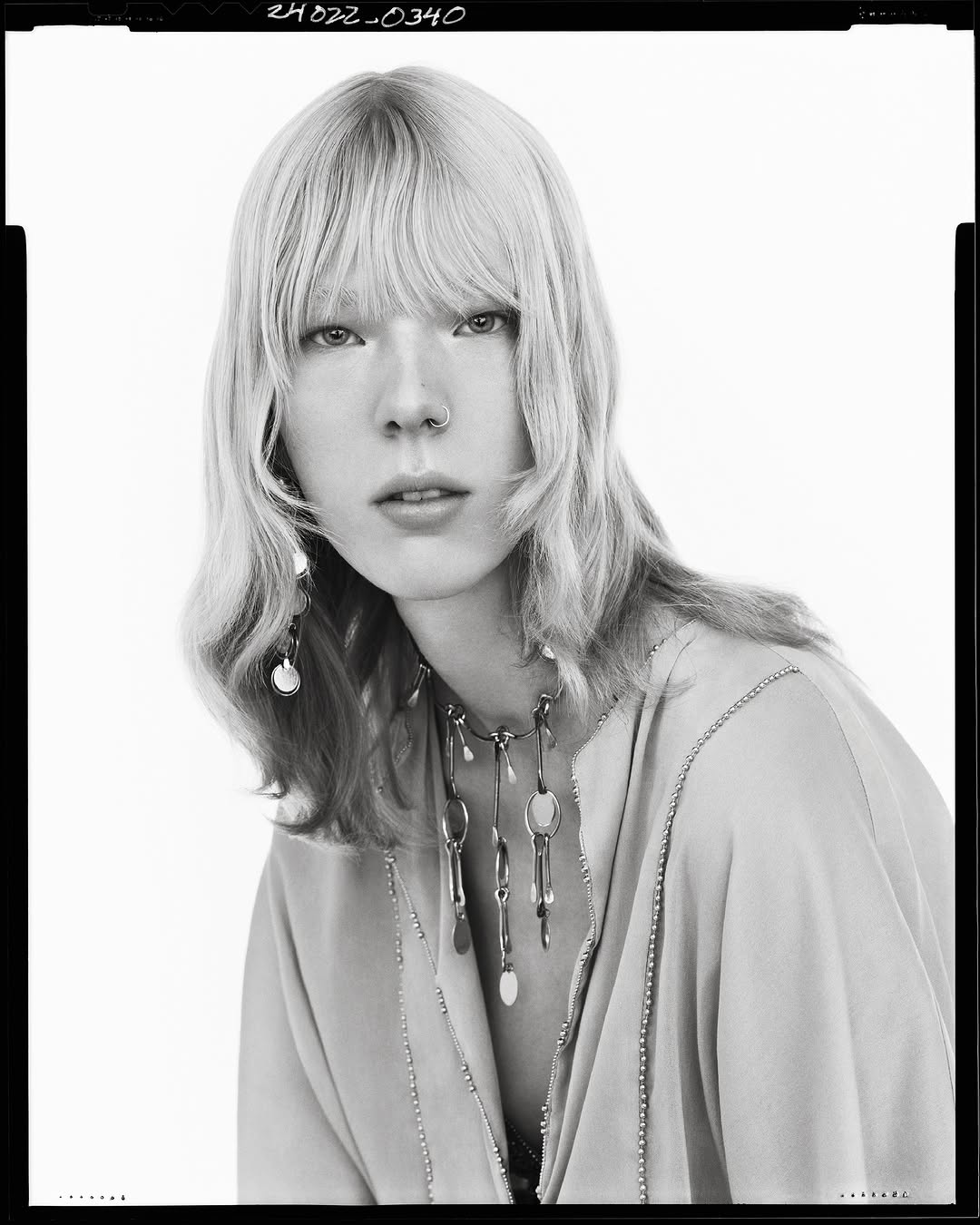 Black and white portrait of a model with blonde hair, bangs, and statement necklace against a seamless background