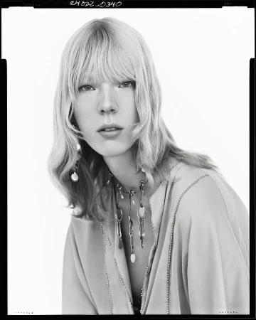 Black and white portrait of a model with blonde hair, bangs, and statement necklace against a seamless background