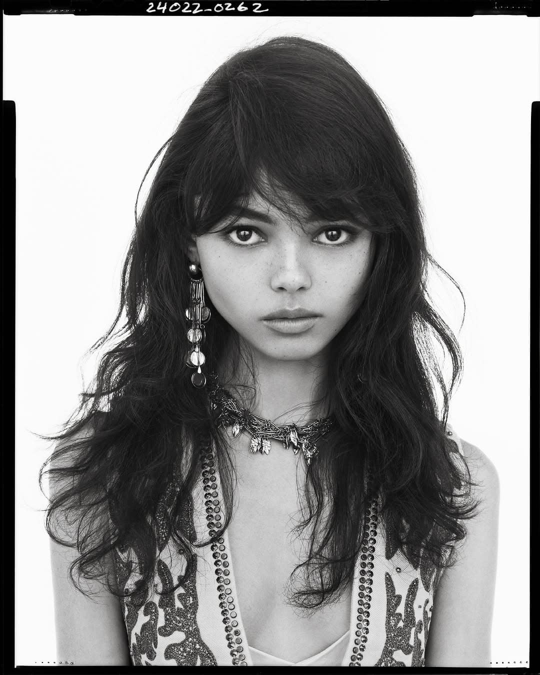 Portrait of a model with long dark hair, statement earrings, and patterned clothing against a plain background.