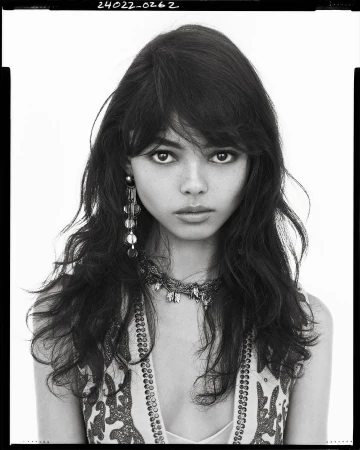 Portrait of a model with long dark hair, statement earrings, and patterned clothing against a plain background.