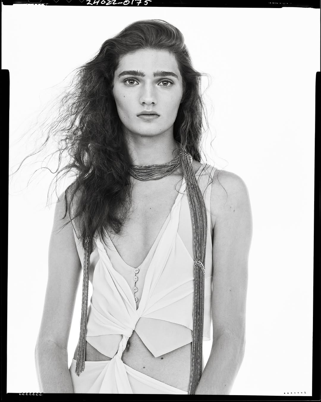 Monochrome portrait of a model with long dark hair wearing a sleeveless top and beaded necklace against a plain white background