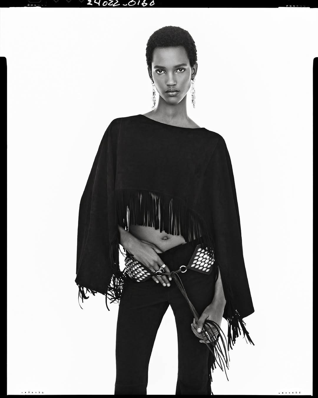 Black and white fashion portrait of a model wearing a fringed top and studded belt against a white seamless background.