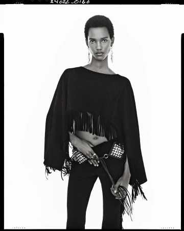 Black and white fashion portrait of a model wearing a fringed top and studded belt against a white seamless background.