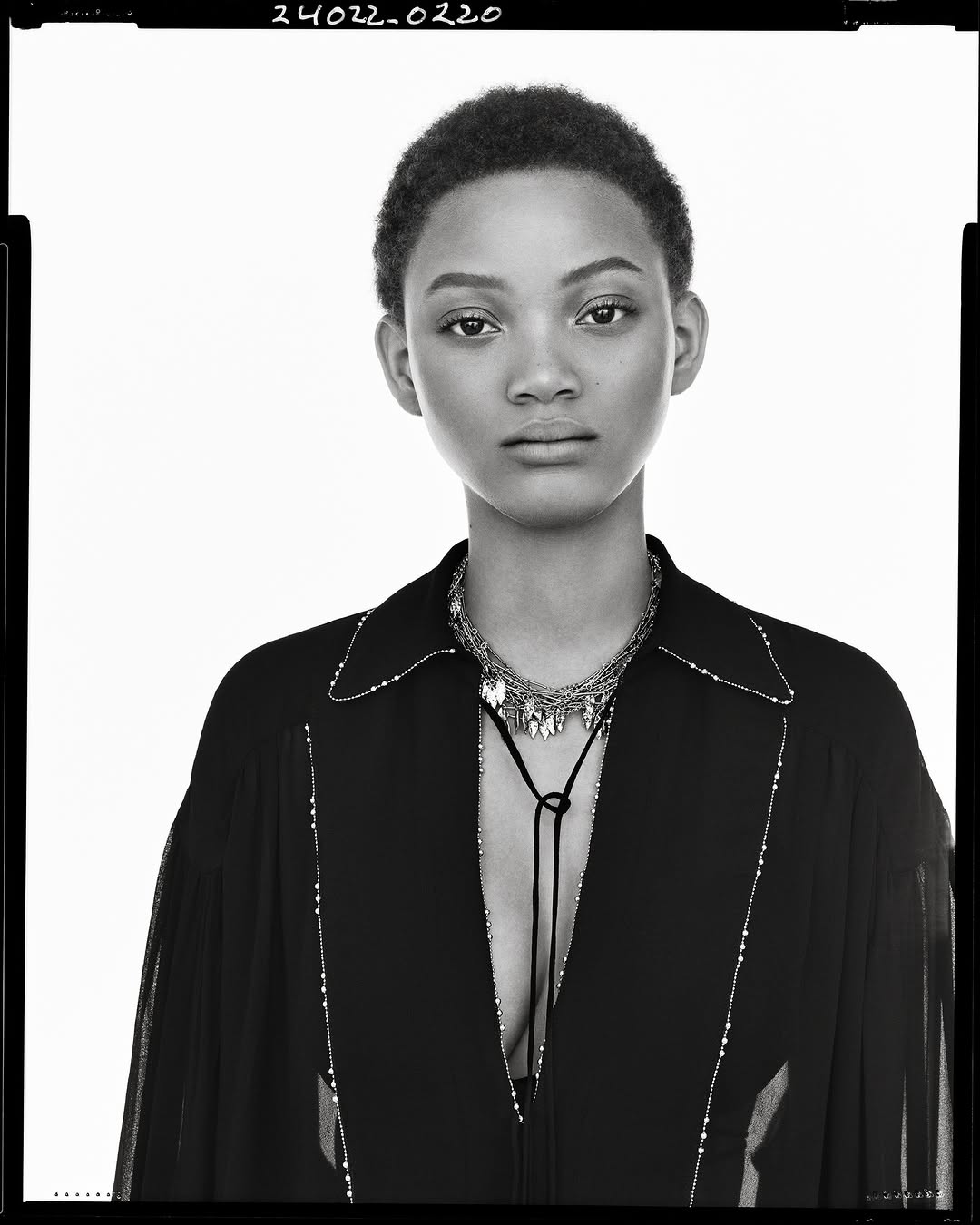 Black and white portrait of a model with short cropped hair wearing a dark collared top and layered necklace against white background