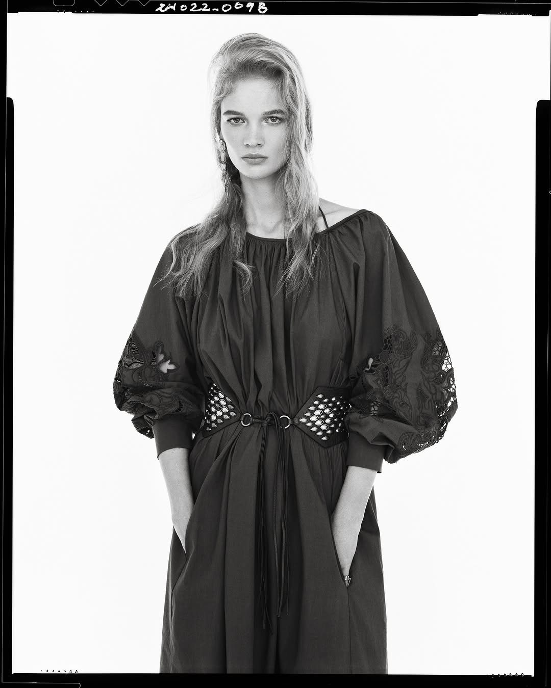 Model wearing dark bishop-sleeve dress with wide grommet belt against white seamless background