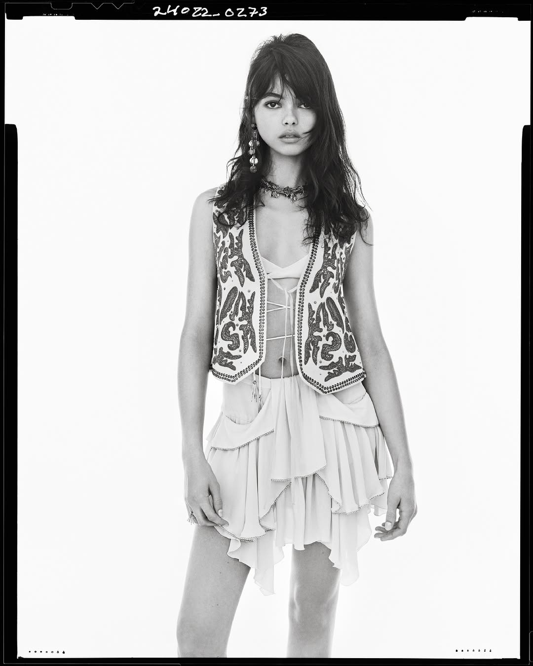 Model in embroidered vest and skirt standing against a plain white studio backdrop with visible film border