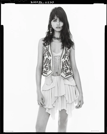 Model in embroidered vest and skirt standing against a plain white studio backdrop with visible film border