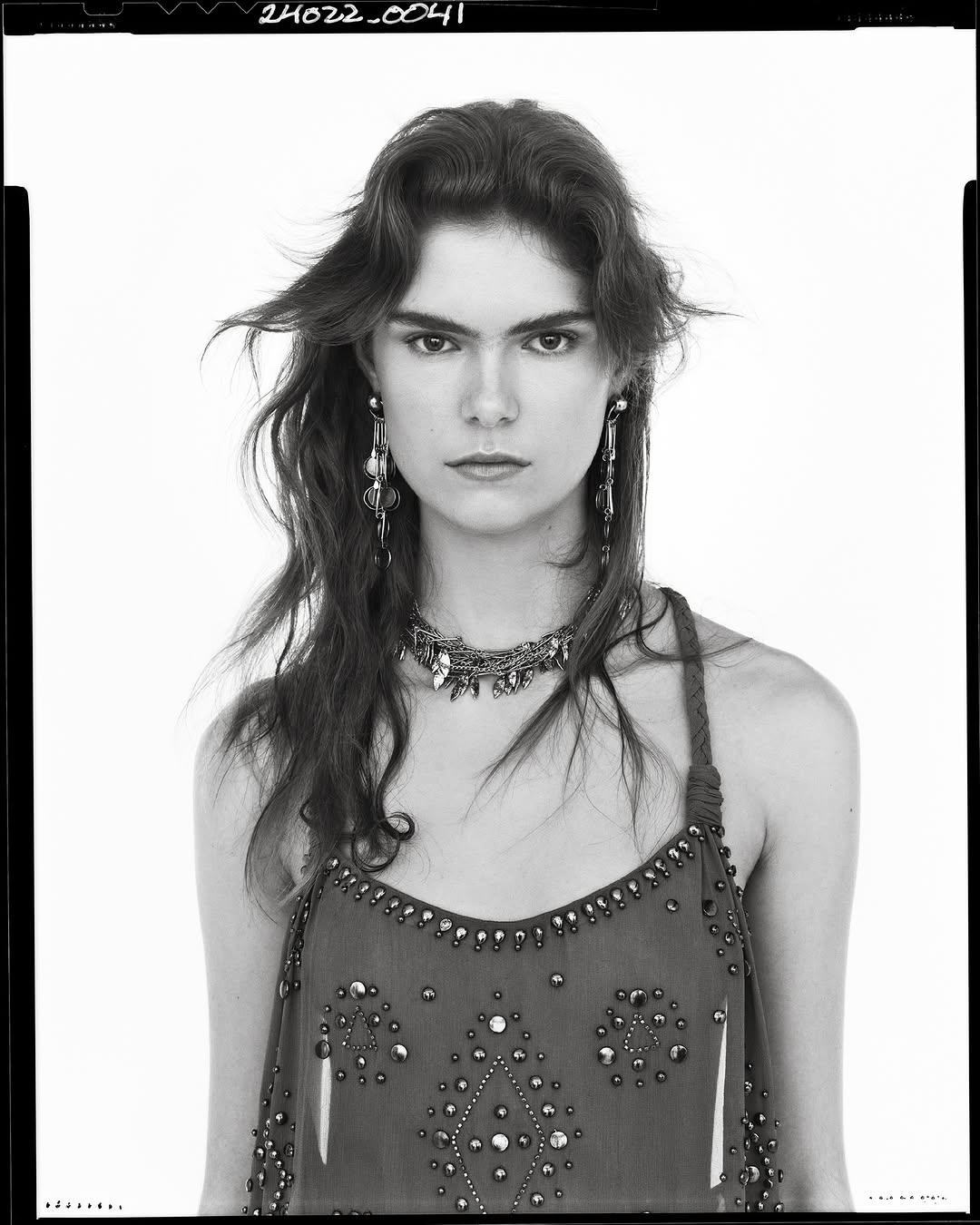 Portrait of a woman wearing a heavily embellished strap dress and ornate jewelry against a white studio background.