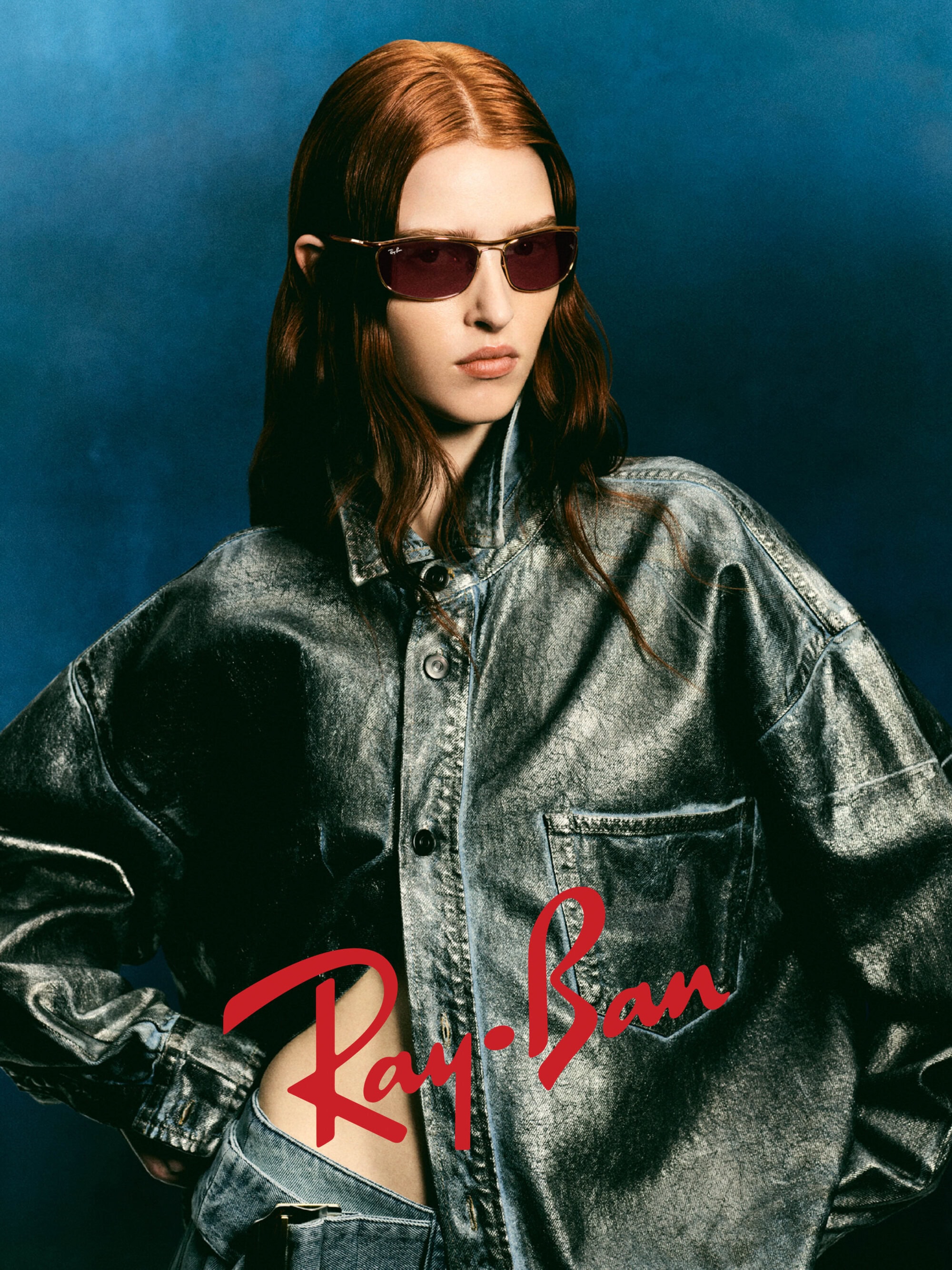 Model with red hair wearing sunglasses and a metallic jacket against a dark blue background with Ray-Ban logo