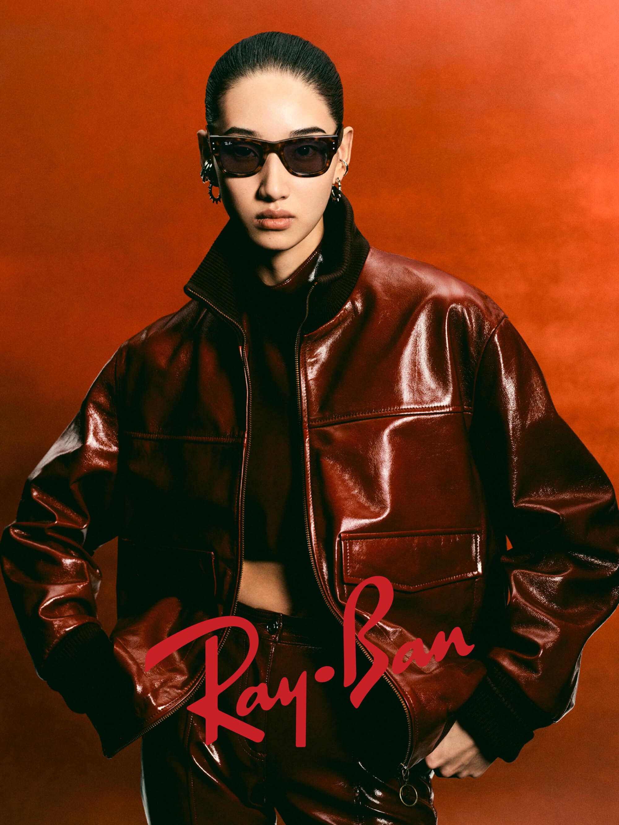 Model wearing dark sunglasses and a glossy brown leather jacket against a rich rust-colored background.