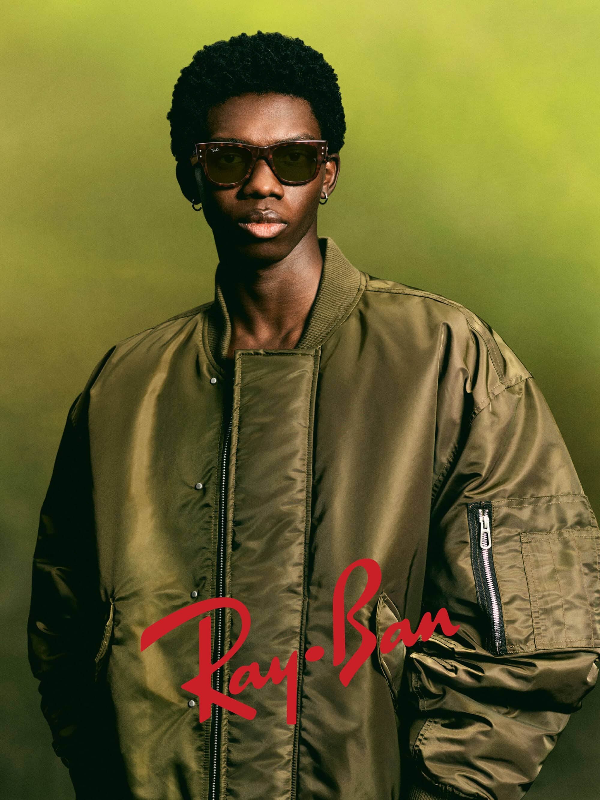 Black man wearing sunglasses and an olive green bomber jacket against a soft gradient background for Ray-Ban