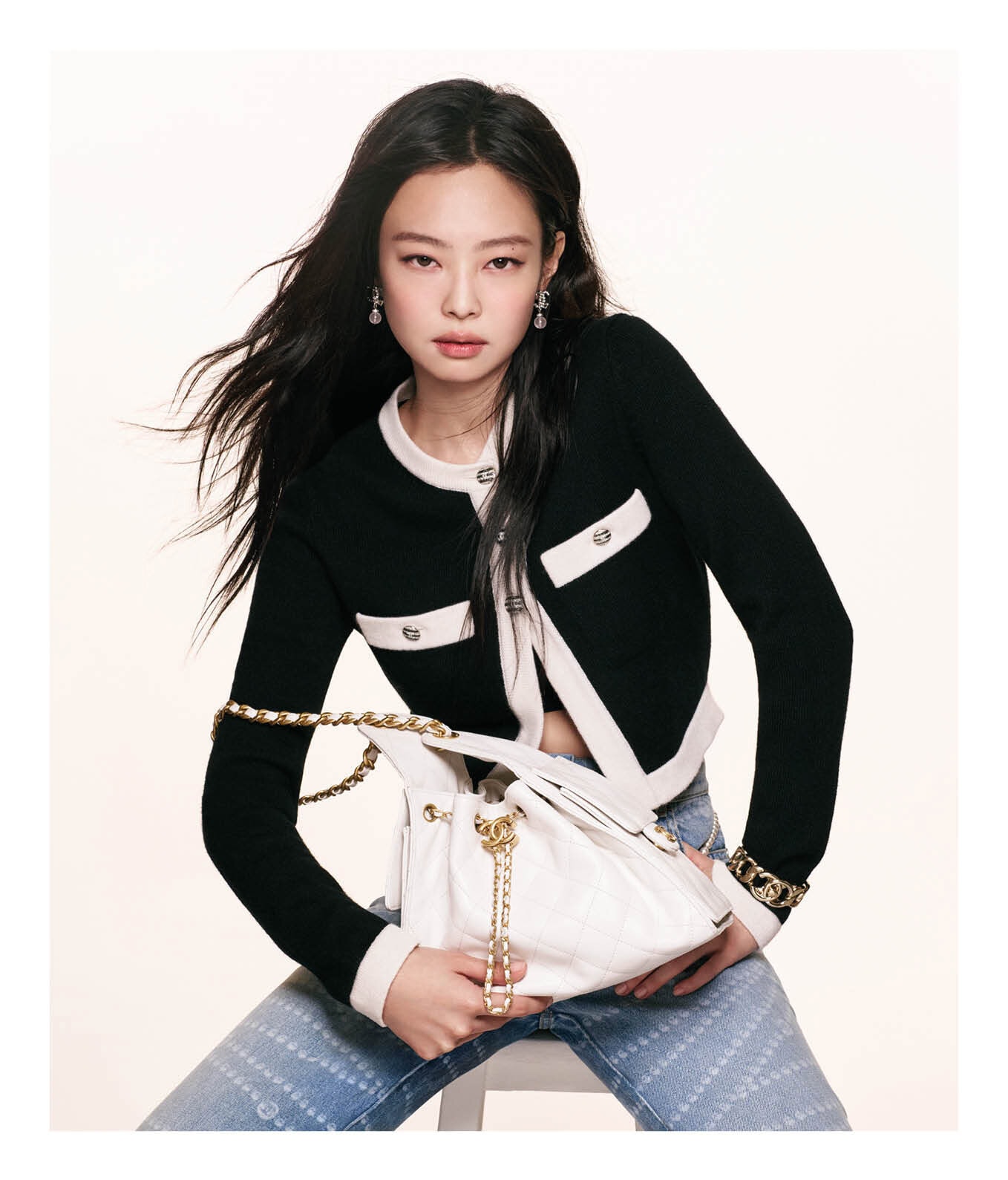 Jennie holding a white quilted handbag wearing a black and white cardigan and patterned jeans against a plain white background