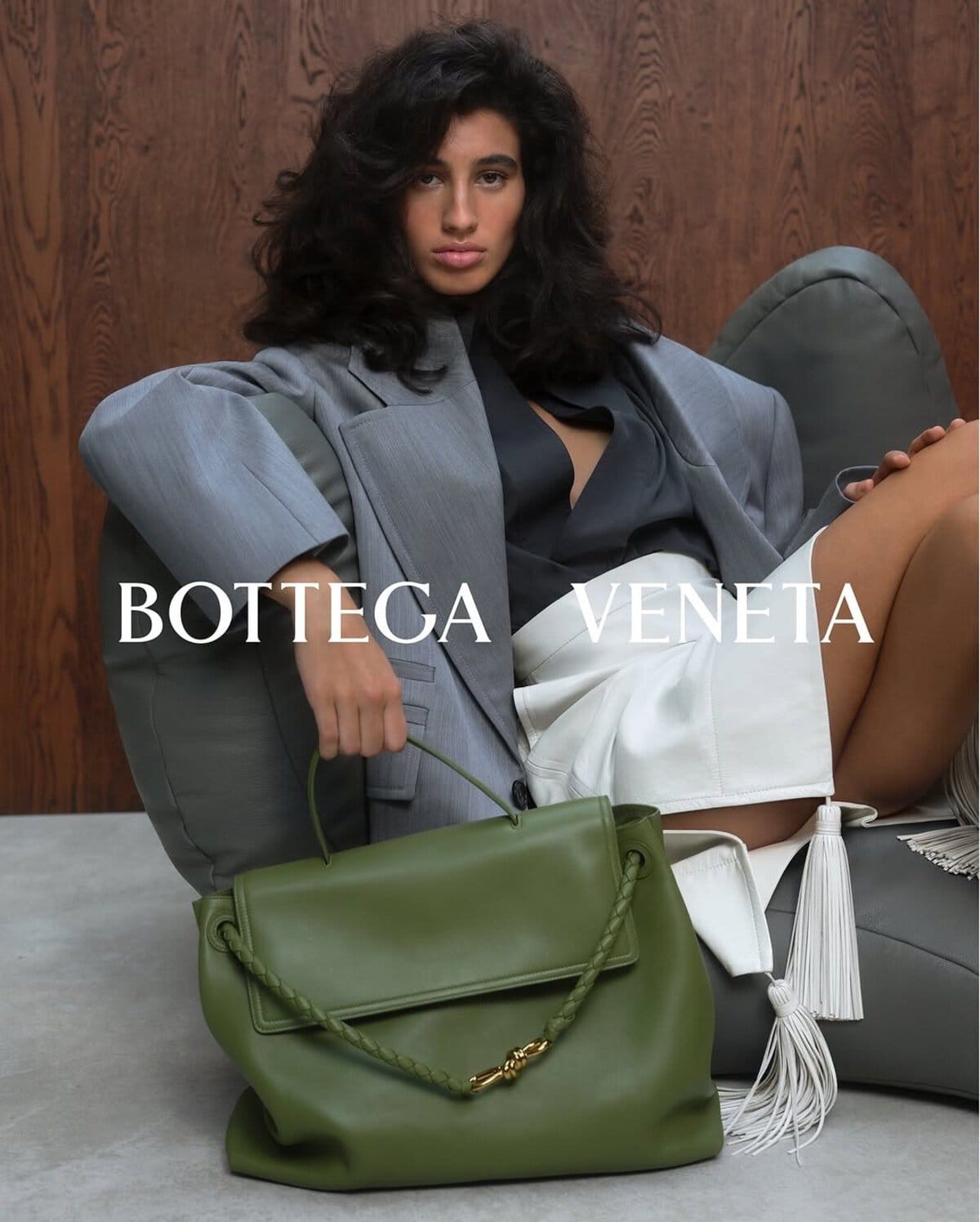 Model seated on a grey lounge chair holding an olive green leather handbag for Bottega Veneta Spring 2025 campaign.