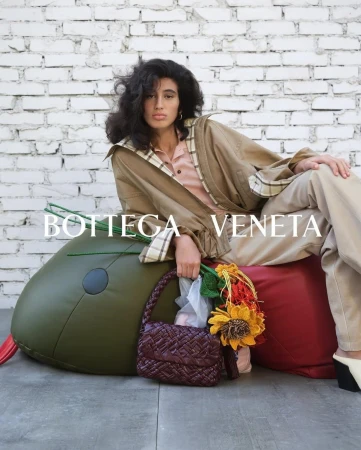 Model seated against a white brick wall holding a woven dark red leather handbag and flowers in front of green and red beanbag furniture.