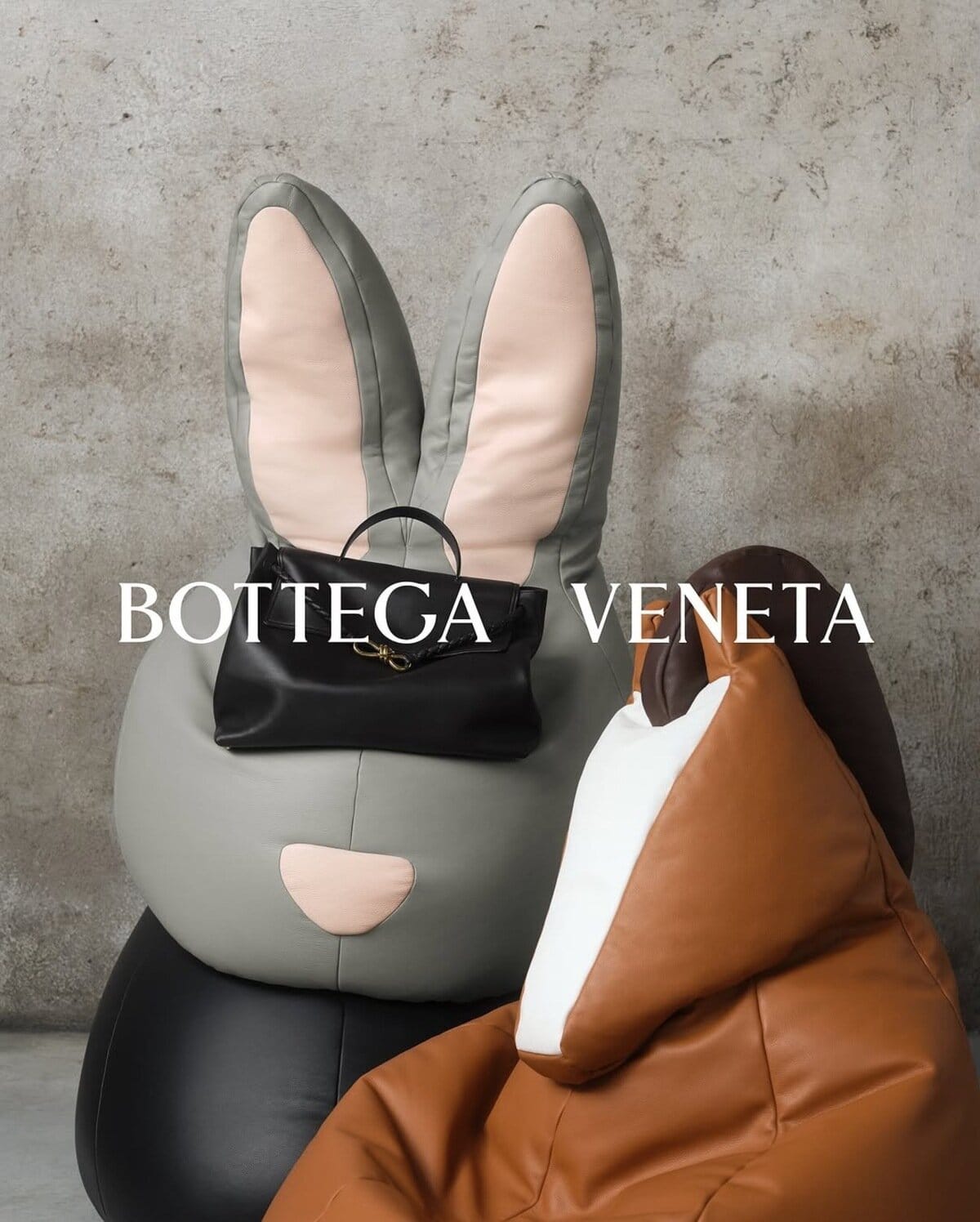 Grey rabbit bean bag chair with pink inner ears and a black leather handbag resting on it against a textured concrete wall.