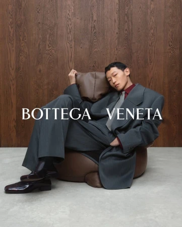 Man seated in brown leather armchair wearing oversized grey suit and tie against wooden paneling background