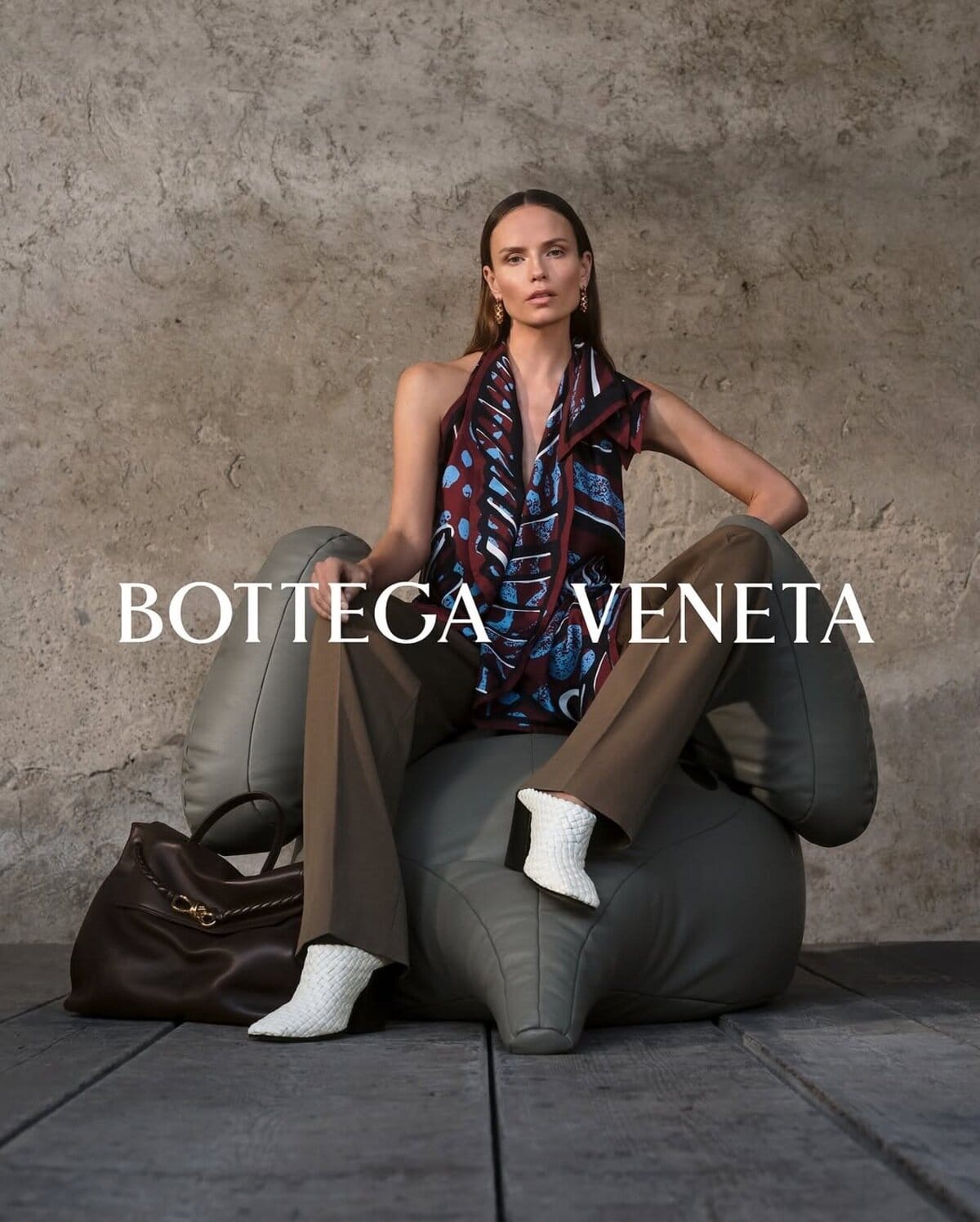 Model seated on a grey leather armchair wearing a patterned halter top and brown trousers with a large dark bag nearby against a textured wall