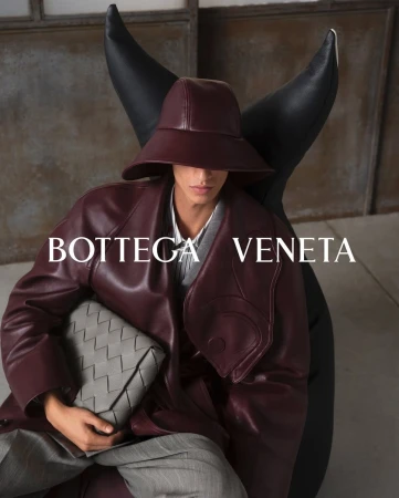 Model seated wearing a burgundy leather coat and hat, holding a grey woven clutch against an industrial background.