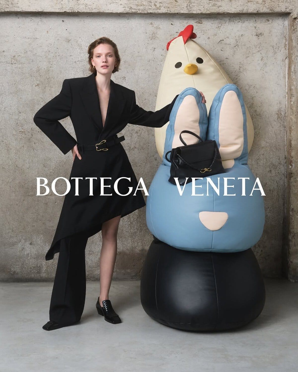 Model in black blazer dress posing beside a whimsical blue and cream sculpture against a weathered concrete wall