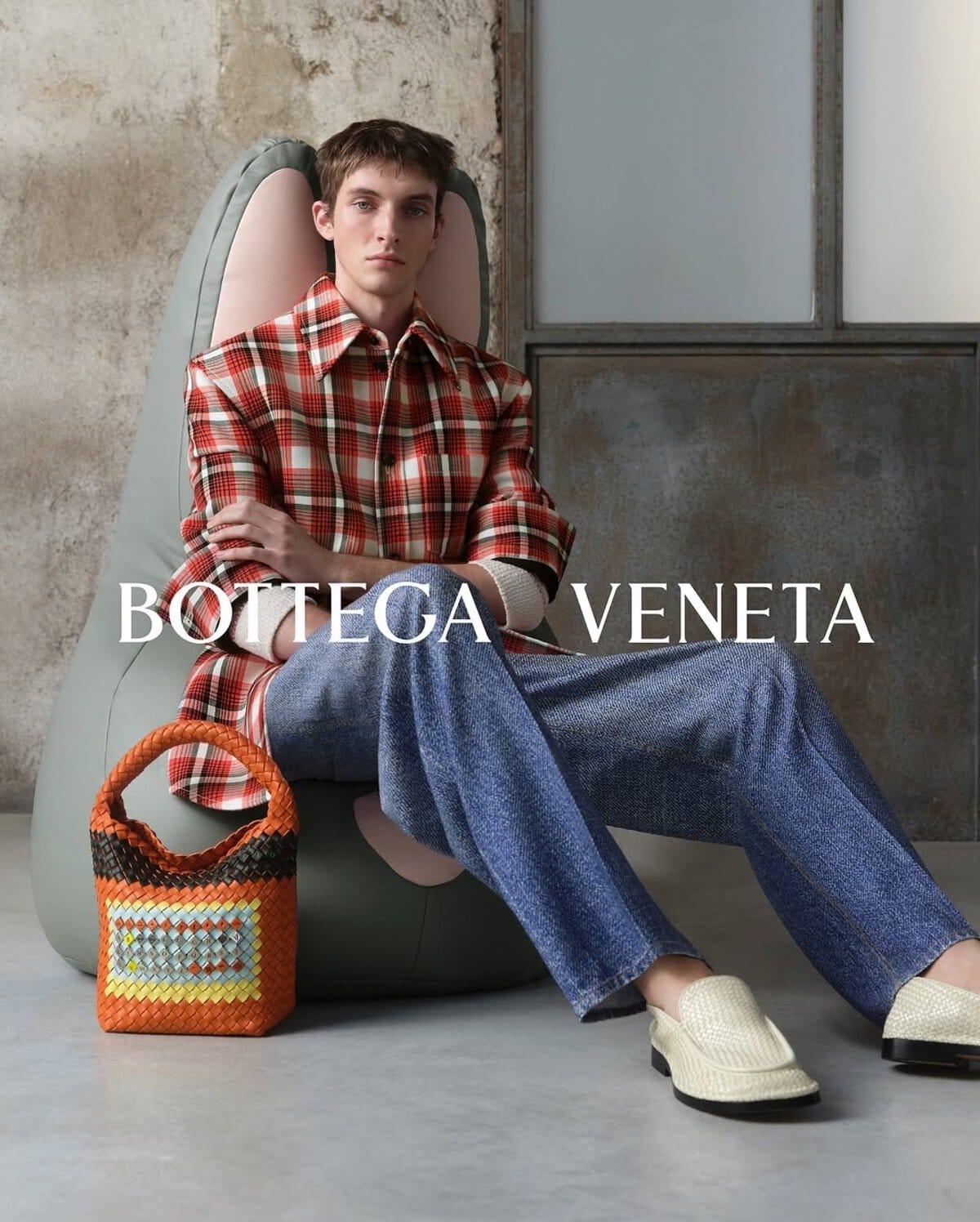 Model seated in an armchair wearing a red plaid shirt and blue jeans with a woven orange handbag beside them