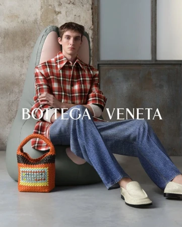 Model seated in an armchair wearing a red plaid shirt and blue jeans with a woven orange handbag beside them