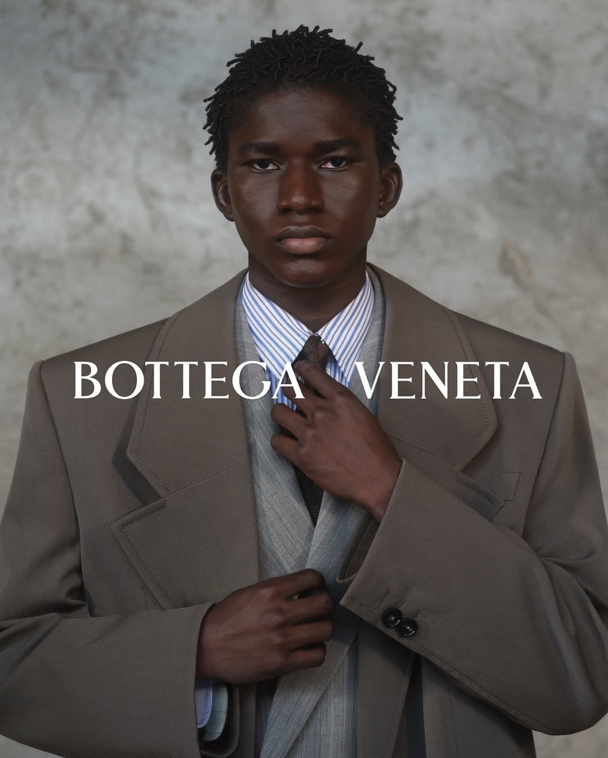 Black man adjusting tie wearing brown suit jacket and striped shirt against textured wall