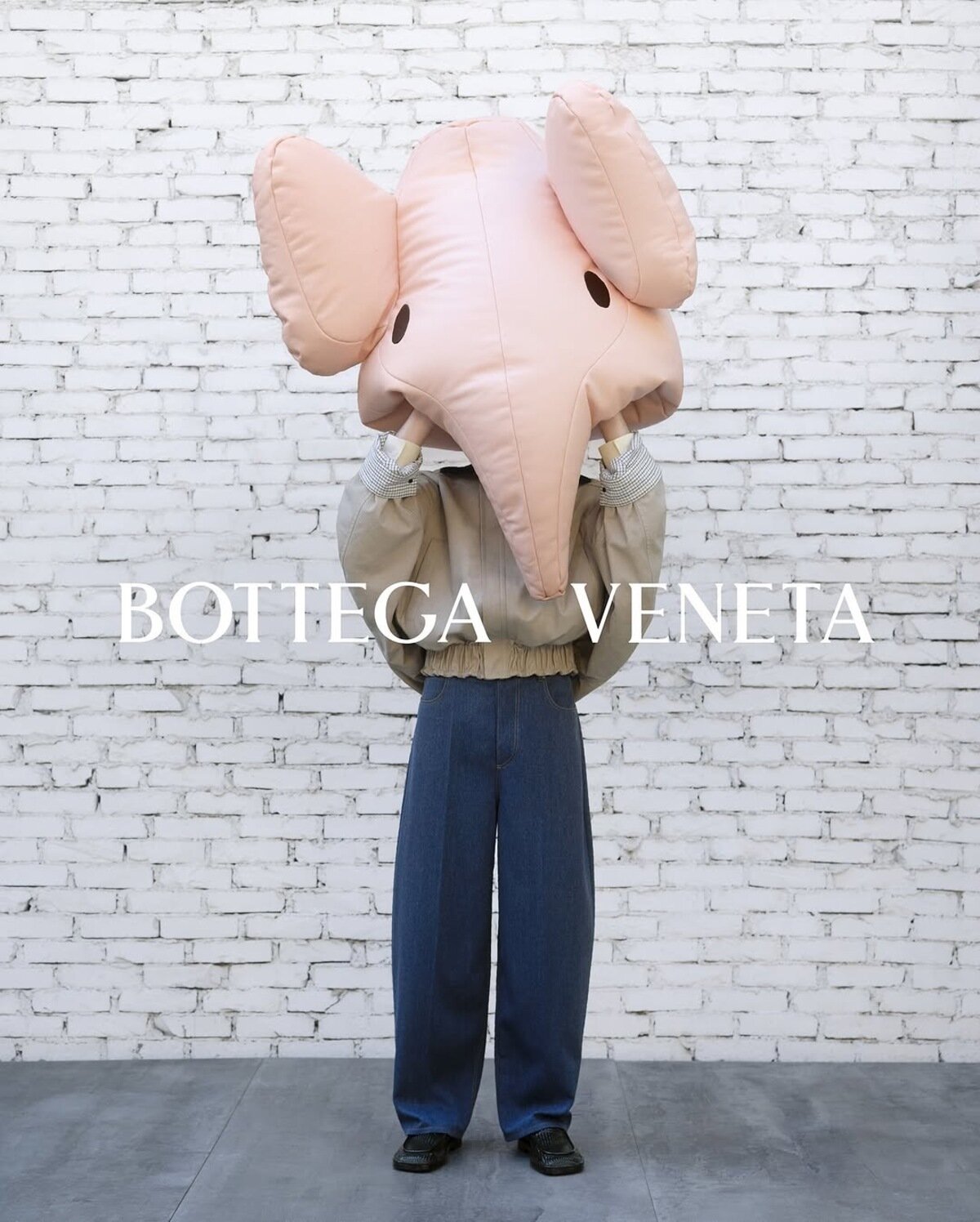 Person holding large pink elephant mask wearing grey jacket and blue jeans in front of white brick wall