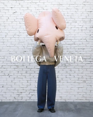 Person holding large pink elephant mask wearing grey jacket and blue jeans in front of white brick wall