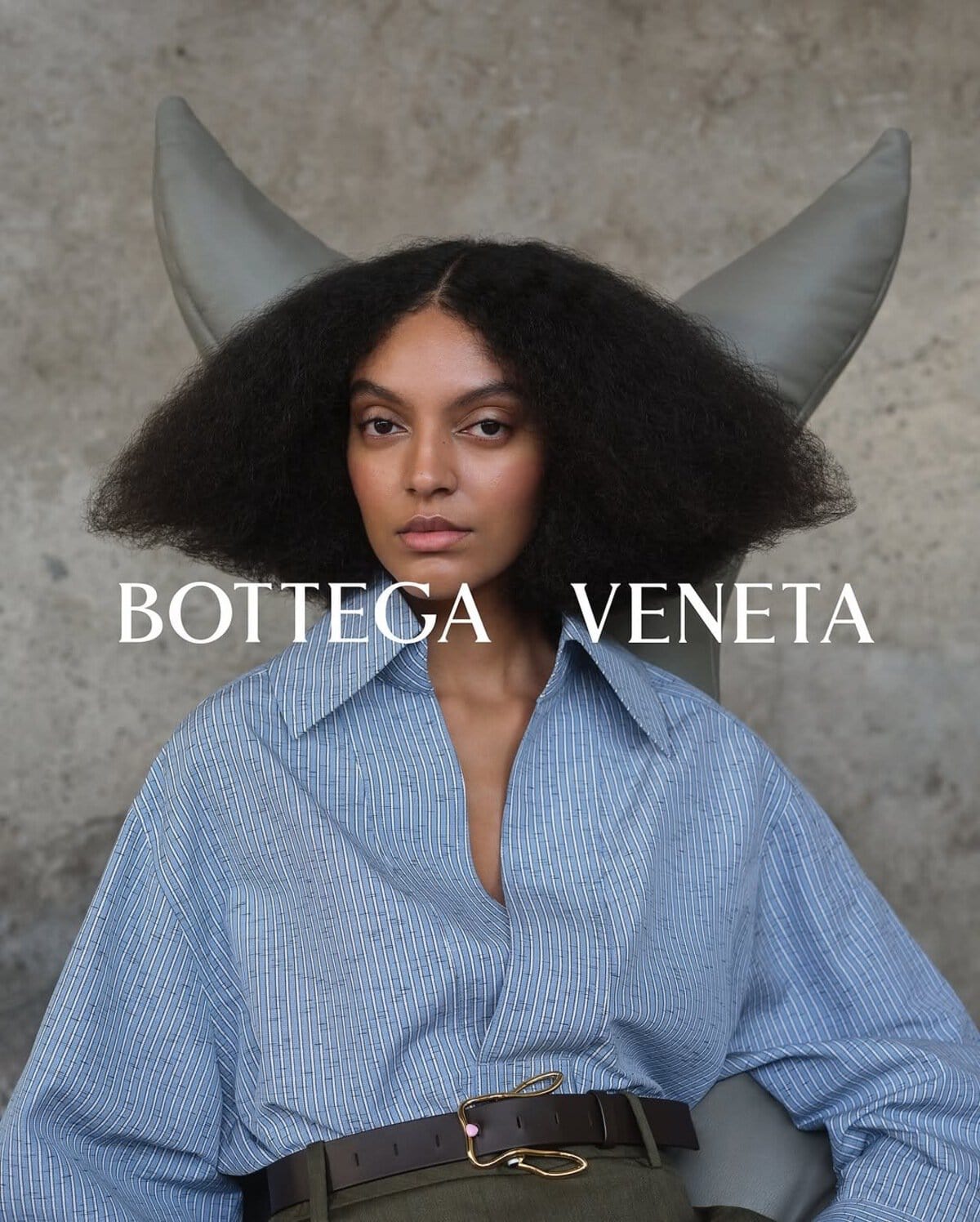 Model with voluminous dark hair wearing a blue button-down shirt and brown leather belt against a textured grey background.