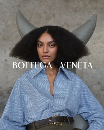 Model with voluminous dark hair wearing a blue button-down shirt and brown leather belt against a textured grey background.