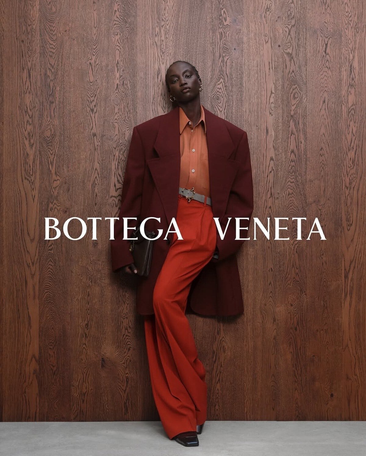 Model wearing an oversized burgundy blazer and red trousers standing against a rich wood panel wall