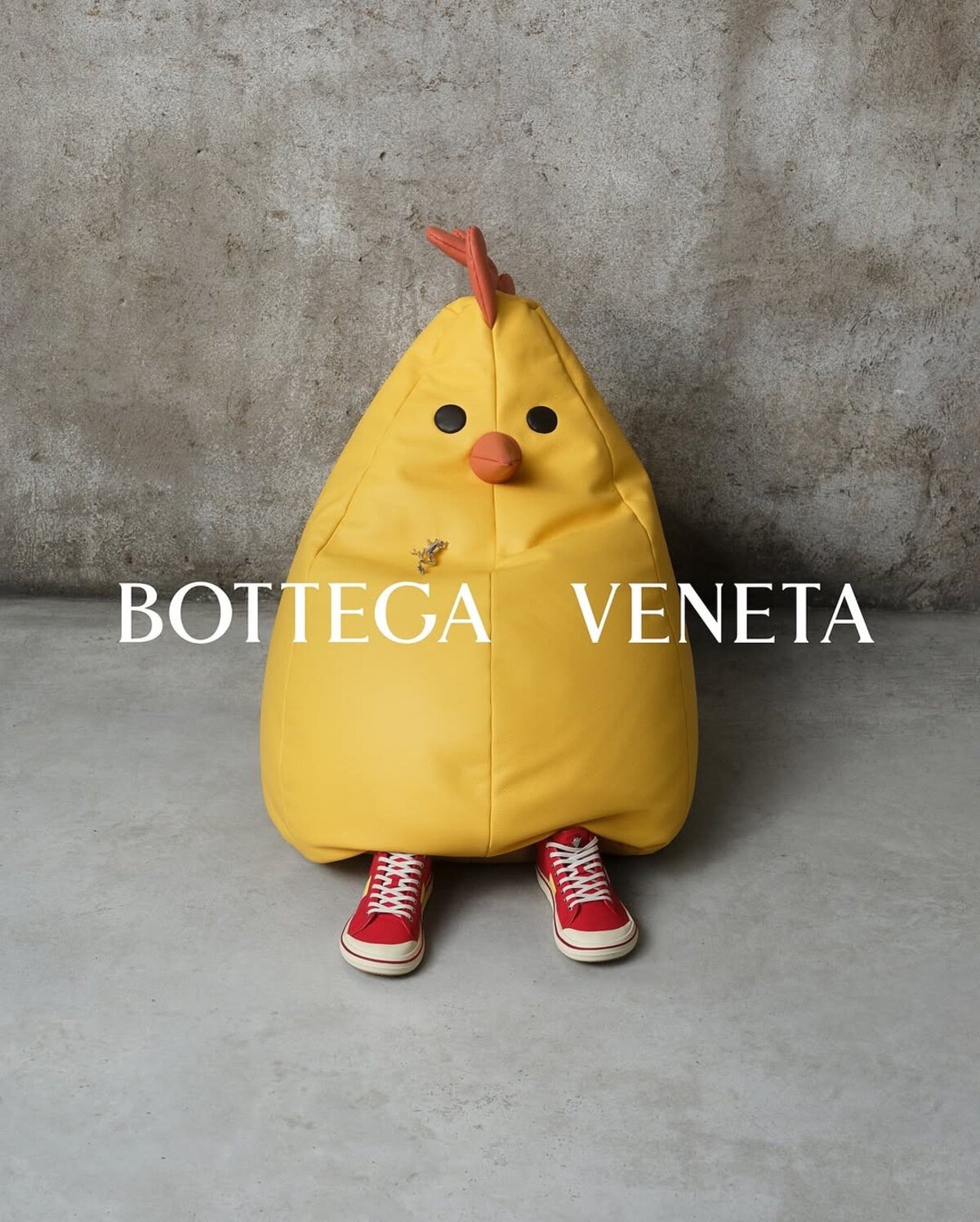 A person wearing a large yellow chicken costume and red sneakers sitting against a textured wall with Bottega Veneta text overlay.