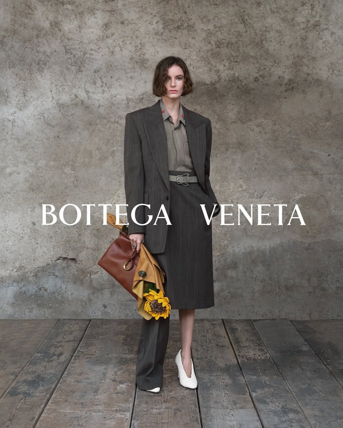 Model wearing dark blazer and skirt holding a leather bag with sunflowers, set against a rustic stone wall.